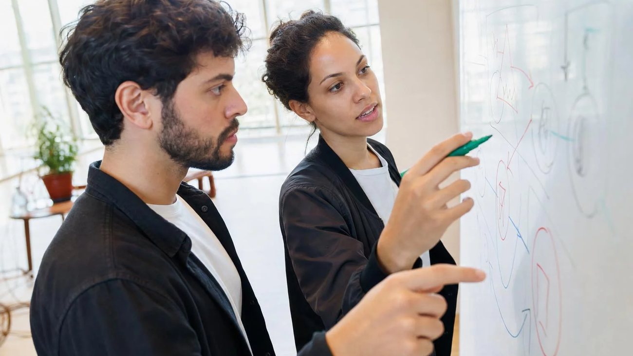 Duas participantes de pé diante do quadro branco, montando um argumento a caneta